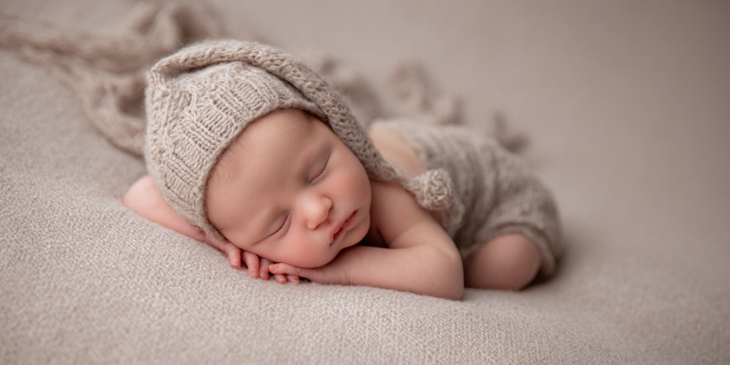 A newborn baby sleeps peacefully in a side-lying pose on a soft beige blanket, wrapped in neutral-toned fabric inside a cozy, softly lit studio setting.