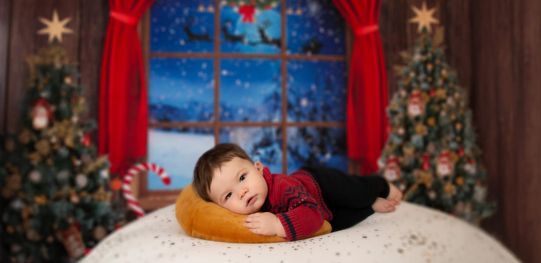 Baby in a red sweater lying on a pillow during a Christmas photoshoot with decorated trees and snowy window backdrop.